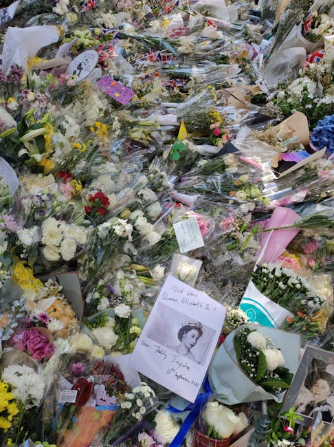 A large memorial display of numerous floral tributes and funeral flowers laid on the ground, featuring a variety of bouquets with roses, carnations, and lilies in colors such as white, pink, purple, a