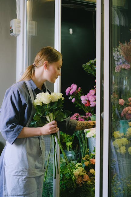 A female florist with light brown hair tied back, dressed in a grey apron over a blue shirt, is selecting a bouquet of white roses with lush green foliage. She is standing indoors near a glass display