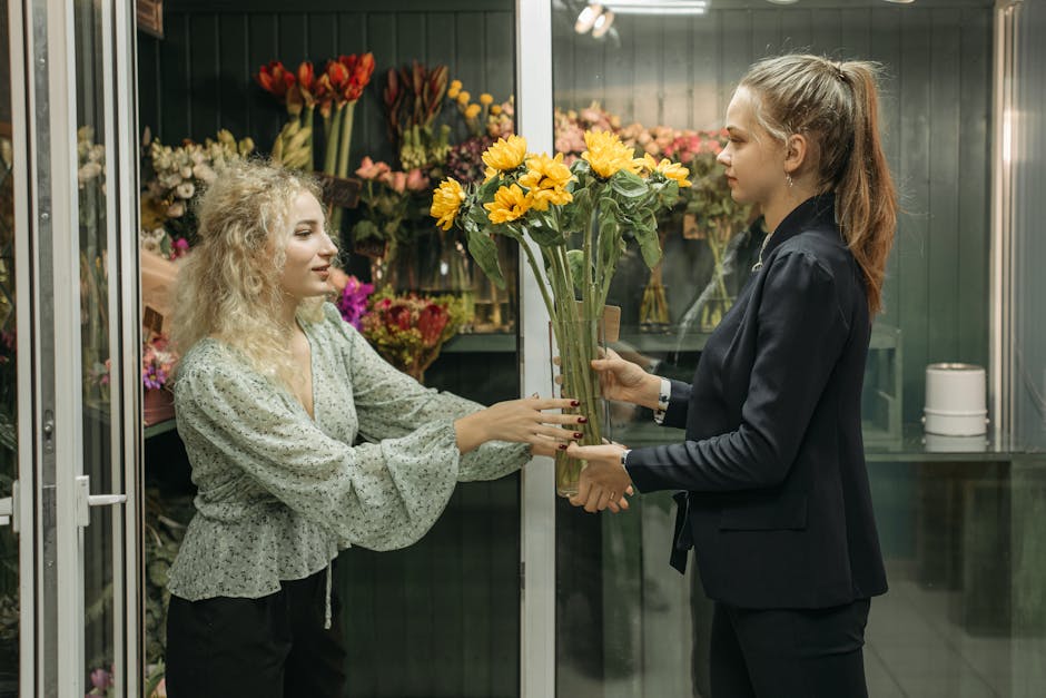 A female florist with curly blonde hair, wearing a light-colored, long-sleeved blouse with a subtle floral pattern, hands a bouquet of vibrant yellow flowers with large, open petals and green stems to