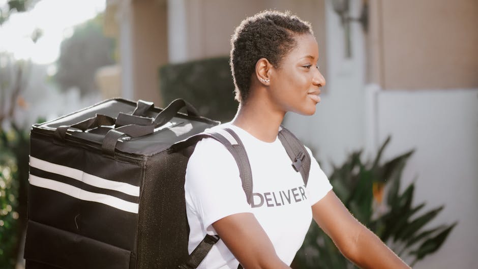A female delivery person with short curly hair and medium skin tone, wearing a white T-shirt with 'DELIVER' printed on it, is outdoors holding a large, black insulated delivery bag with white stripes.